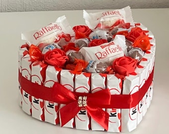 May include: A heart-shaped arrangement of chocolate bars, wrapped in white and red foil, with red roses and assorted chocolates. The arrangement is tied with a red ribbon and a sparkly bow.
