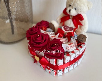 Könnte beinhalten: Eine herzförmige Anordnung von Schokoladentafeln, verziert mit roten Rosen und einem weißen Teddybär, der eine rote Schleife und ein Herz mit der Aufschrift "Ich liebe dich" hält.