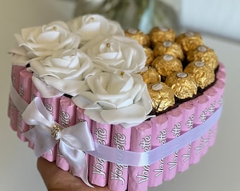 Könnte beinhalten: Eine herzförmige Geschenkbox mit weißen Rosen und goldverpackten Pralinen. Die Box ist mit rosa Schokoriegeln und einem weißen Band mit Strass verziert. Eine einzigartige und festliche Geschenkidee.
