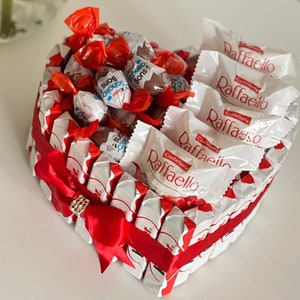 May include: A heart-shaped arrangement of chocolate candies wrapped in red and white wrappers. The candies are arranged in a heart shape and tied with a red ribbon. The candies are labelled "Raffaello" and "Baci Perugina".