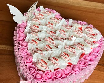 Pode incluir: Uma caixa de presente em forma de coração cheia de doces Raffaello, rodeada por rosas cor-de-rosa e um laço decorativo. Uma borboleta branca adiciona um toque delicado. Um presente charmoso para qualquer ocasião.