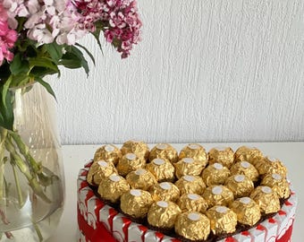 Könnte beinhalten: Eine herzförmige Anordnung von Schokoladenbonbons, die in goldene Folie verpackt sind, umgeben von kleinen Schokoladentafeln mit rotem Band und einem goldenen Herz-Anhänger.