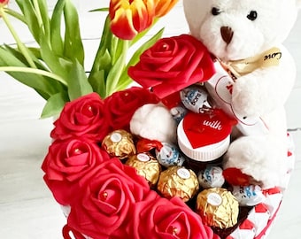 Könnte beinhalten: Eine herzförmige Anordnung aus roten Rosen und Schokoladenbonbons, mit einem weißen Teddybär, der eine Schokoladentafel mit der Aufschrift "Ich liebe dich" hält.