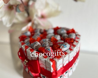 May include: A heart-shaped box filled with chocolate candies, wrapped in white and red foil. The box is decorated with a red ribbon and a gold charm. The box is surrounded by white flowers.