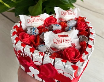 Könnte beinhalten: Eine herzförmige Schachtel gefüllt mit verschiedenen Schokoladen und roten Rosen. Die Schachtel besteht aus weißen Schokoladenverpackungen und ist mit einem roten Band verziert. Die Schokoladen umfassen Raffaello, Ferrero Rocher und andere verschiedene Schokoladen.