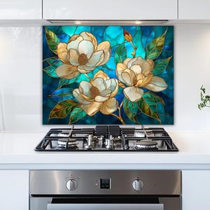 Stained glass style backsplash featuring three cream magnolias on a vibrant turquoise background, installed behind a silver gas range in a white modern kitchen with stainless steel appliances and minimalist cabinetry.