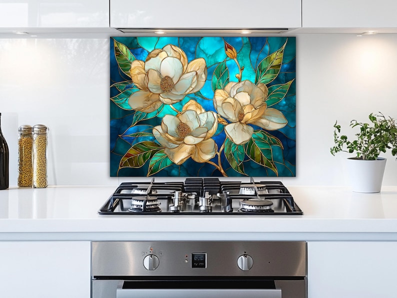Stained glass style backsplash featuring three cream magnolias on a vibrant turquoise background, installed behind a silver gas range in a white modern kitchen with stainless steel appliances and minimalist cabinetry.