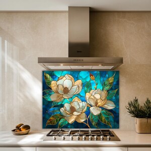 Stained glass style backsplash featuring three cream magnolias on a turquoise background, installed behind a silver gas range in a modern tan stone-tiled kitchen with a minimalist steel vent hood.