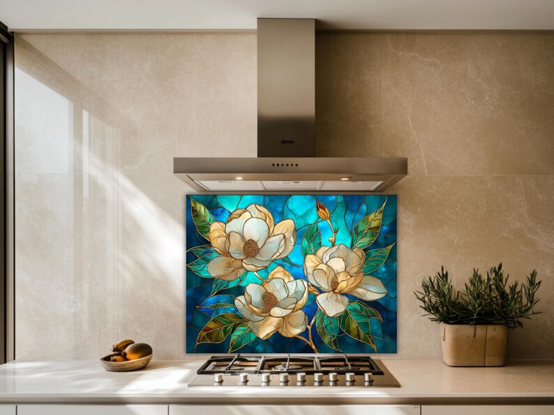 Stained glass style backsplash featuring three cream magnolias on a turquoise background, installed behind a silver gas range in a modern tan stone-tiled kitchen with a minimalist steel vent hood.