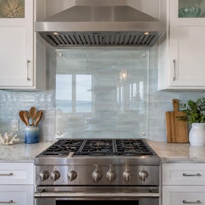 May include: A stainless steel gas range with a large oven, six burners, and a glass backsplash panel. The kitchen features white cabinetry, a stainless steel range hood, a blue ombre utensil holder with wooden spoons, and a wooden cutting board.