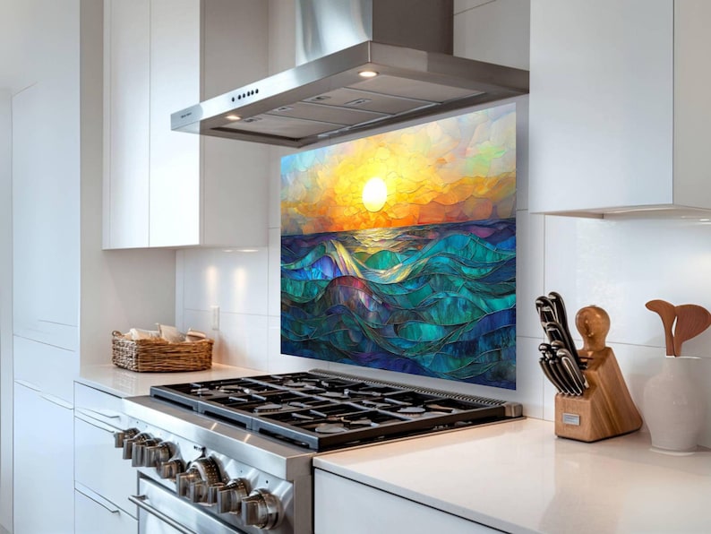 A stained-glass style backsplash featuring an ocean sunset with rolling waves is displayed in a modern white kitchen. It sits behind a gas cooktop, flanked by a woven basket and a knife block.