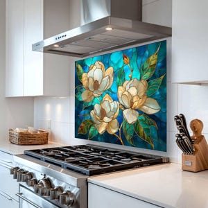 Stained glass style backsplash featuring three cream magnolias on a vibrant turquoise background, installed behind a silver gas range in a white modern kitchen with stainless steel appliances and a minimalist range hood.