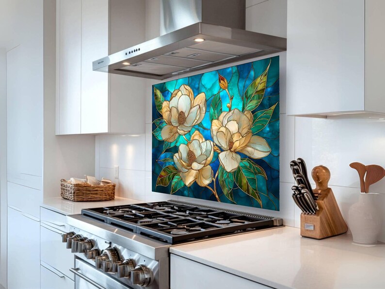 Stained glass style backsplash featuring three cream magnolias on a vibrant turquoise background, installed behind a silver gas range in a white modern kitchen with stainless steel appliances and a minimalist range hood.