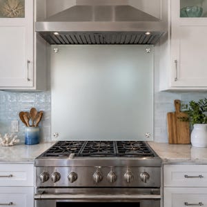 May include: A rectangular frosted glass kitchen backsplash panel mounted with four silver circular hardware fasteners, positioned directly behind a stainless steel gas range stove and beneath a matching stainless steel range hood vent.