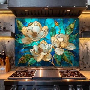 Stained glass style backsplash featuring three large cream-colored magnolias on a vibrant blue and turquoise background, installed behind a black gas range in an industrial-style kitchen with dark concrete walls and warm LED lighting.
