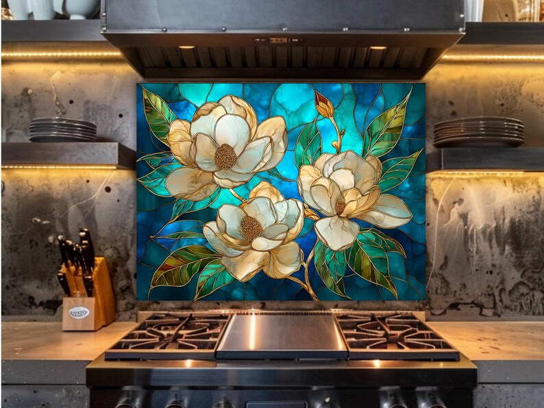 Stained glass style backsplash featuring three large cream-colored magnolias on a vibrant blue and turquoise background, installed behind a black gas range in an industrial-style kitchen with dark concrete walls and warm LED lighting.