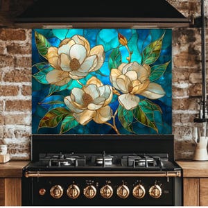 Stained glass style backsplash featuring three large cream-colored magnolias with green leaves on a vibrant blue and turquoise background, installed behind a black gas range in a rustic brick kitchen.