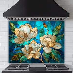 Stained glass style backsplash featuring three large cream-colored magnolias with green leaves on a vibrant blue and turquoise background, installed behind a black gas range in a white subway-tiled kitchen.