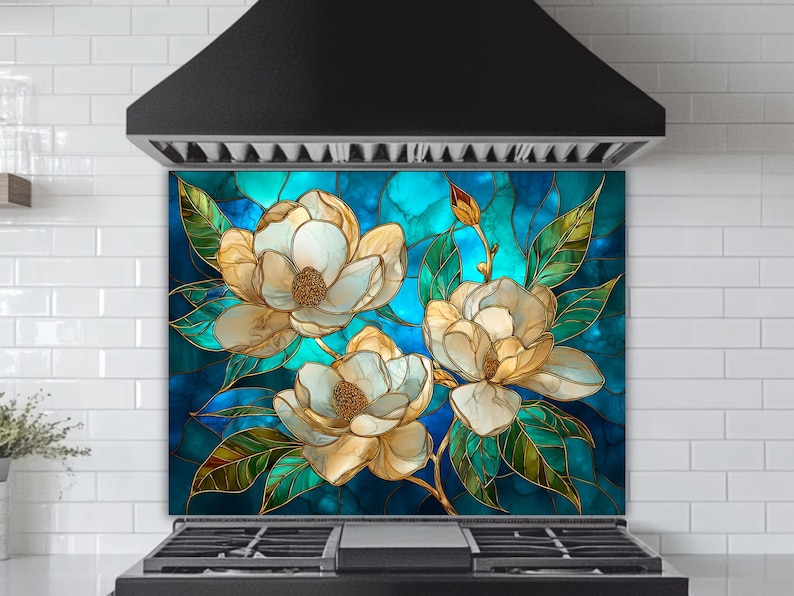 Stained glass style backsplash featuring three large cream-colored magnolias with green leaves on a vibrant blue and turquoise background, installed behind a black gas range in a white subway-tiled kitchen.