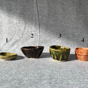 May include: Four small, handcrafted ceramic bowls in varying colors and textures. The bowls include a shallow green dish, a dark brown bowl, a green bowl with textured sides, and a reddish-brown bowl with etched text.