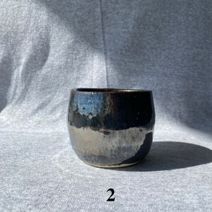 May include: A handcrafted ceramic tumbler with a dark, glossy finish. The cup has a rounded shape and a textured surface, with a metallic sheen. The number "2" is printed in white on the bottom.
