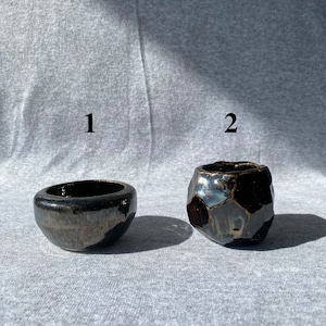 May include: Two small, dark ceramic bowls. One is round with a smooth surface, the other is faceted with a textured surface. Both have a glossy, metallic finish, reflecting light. The bowls are likely for decorative or functional use.