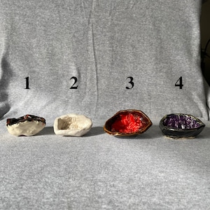 May include: Four small, handmade ceramic bowls in a row. The bowls are various colors, including white, red, and purple. Each bowl has a unique shape and texture, suggesting they are individually crafted. The bowls are displayed on a gray surface.