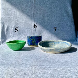 May include: Three handmade ceramic bowls in varying shapes and colors. A small green bowl, a blue bowl with a textured surface, and a light blue bowl with a wavy rim. The bowls are displayed on a gray surface.
