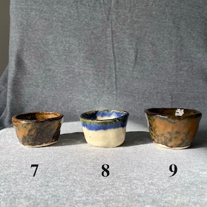 May include: Three small, handmade ceramic bowls with textured surfaces. The bowls are numbered 7, 8, and 9. Bowl 7 is brown and black, bowl 8 is white with a blue rim, and bowl 9 is brown and orange.