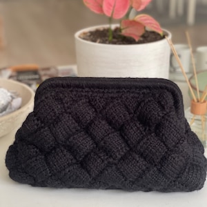 Puede incluir: Un bolso de mano negro de crochet con un diseño tejido. El bolso tiene una forma redondeada, ligeramente curvada y un cierre de broche.