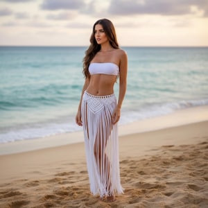 Puede incluir: Un top blanco sin tirantes y una falda de ganchillo blanca con flecos. La falda tiene una cintura alta y un patrón decorativo en la parte superior. El atuendo se usa en una playa de arena con el océano de fondo.