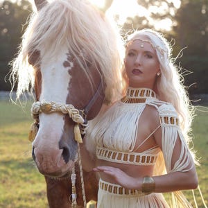 Puede incluir: Una mujer con un atuendo de flecos color crema está de pie junto a un caballo con una melena color crema. La mujer lleva un top y una falda a juego con detalles dorados. El caballo tiene una melena color crema y una brida decorativa crema y dorada.
