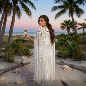 Op de afbeelding: Een witte macramé cape met franjes. De cape wordt gedragen op een strand met palmbomen en een zonsondergang op de achtergrond. De persoon houdt een glas champagne vast.