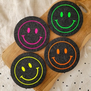 Könnte beinhalten: Vier runde Filzuntersetzer mit schwarzem Hintergrund und einem Neon-Smiley-Gesichtsdesign. Die Untersetzer sind grün, pink, orange und gelb.