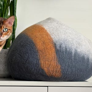 May include: A dome-shaped felted wool pet cave featuring a color-blocked design with sections of dark charcoal gray, a vibrant burnt orange stripe, and a light heather gray top. The structure has a soft, textured surface and a rounded silhouette.