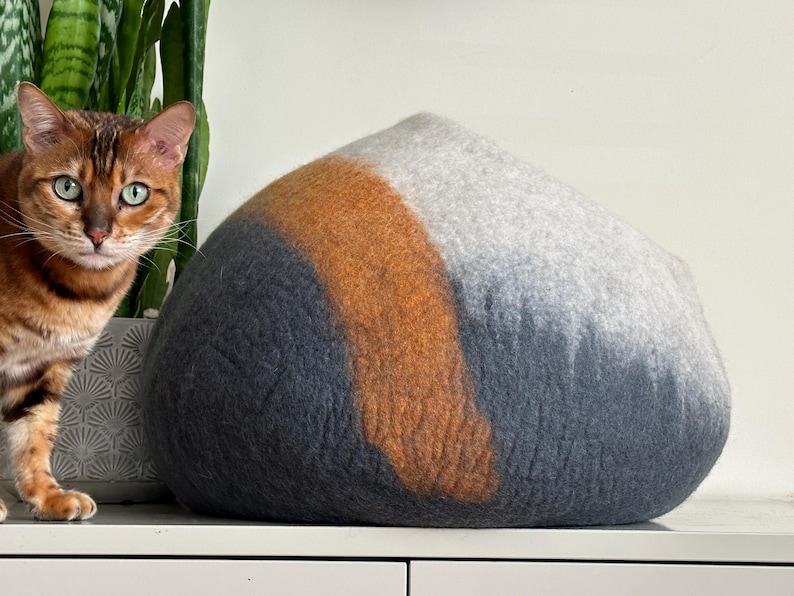 May include: A dome-shaped felted wool pet cave featuring a color-blocked design with sections of dark charcoal gray, a vibrant burnt orange stripe, and a light heather gray top. The structure has a soft, textured surface and a rounded silhouette.