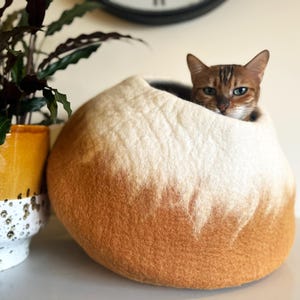 May include: A round, felted wool cat cave featuring an ombre color gradient that transitions from a warm, deep orange at the base to a soft, creamy white at the top. A brown tabby cat is resting inside the circular opening of the pet bed.
