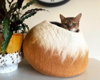 May include: A round, felted wool cat cave featuring an ombre colour gradient that transitions from a creamy white top to a warm, earthy brown base. A brown tabby cat is resting inside the opening of the soft, textured pet bed.