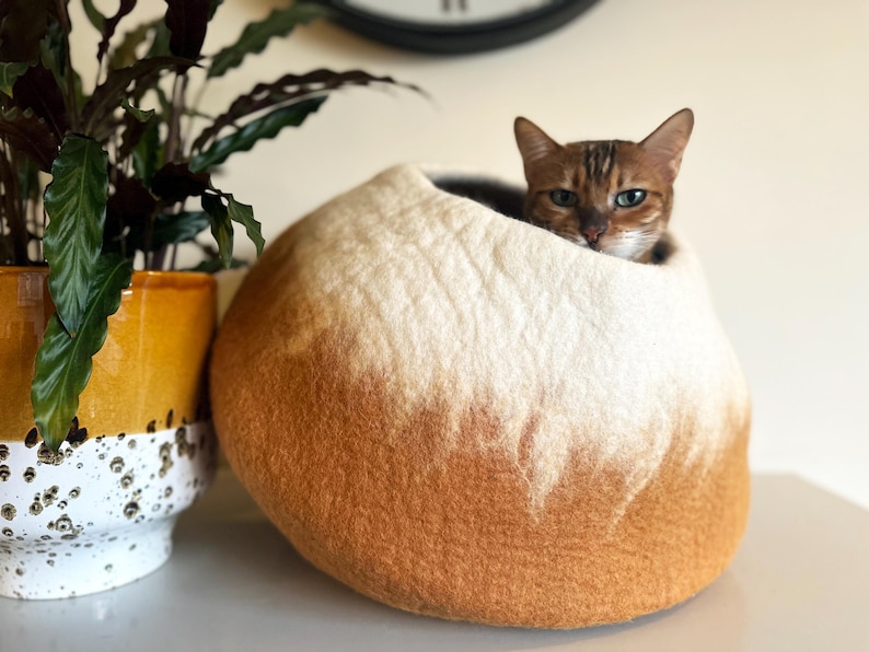 May include: A round, felted wool cat cave featuring an ombre color gradient that transitions from a warm, deep orange at the base to a soft, creamy white at the top. A brown tabby cat is resting inside the circular opening of the pet bed.