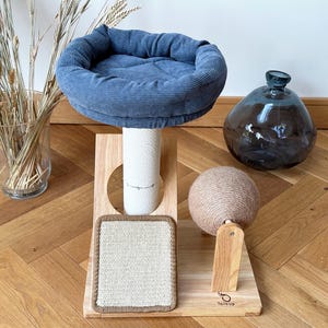May include: A wooden cat tree with a blue corduroy bed, a sisal scratching post, a sisal ball, and a scratching pad. The cat tree is on a wooden floor.