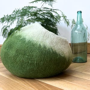 May include: A rounded, felted wool pet cave featuring an ombre color gradient that transitions from a deep forest green at the base to a soft cream color at the top. The structure has a smooth, textured surface and a wide, open circular entrance.