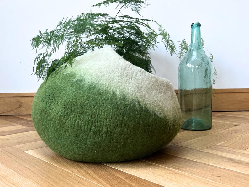 May include: A rounded, felted wool pet cave featuring an ombre color gradient that transitions from a deep forest green at the base to a soft cream color at the top. The structure has a smooth, textured surface and a wide, open circular entrance.