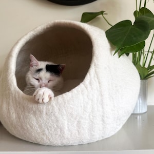 May include: A round, off-white felted wool cat cave with a large circular opening. Inside the cave, a white cat with black markings on its head rests with one paw extended over the edge of the opening, showing its claws.