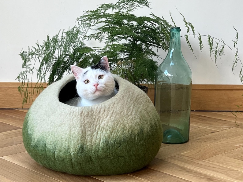 May include: A round, felted wool pet cave featuring an ombre color gradient that transitions from a light cream top to a deep forest green base. A white cat with black markings on its head is resting inside the circular opening of the structure.
