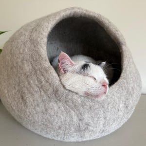 May include: A light gray, rounded, felted wool pet cave with a large circular opening. Inside the soft, textured structure, a white cat with black markings on its head is sleeping peacefully with its eyes closed and pink nose visible.