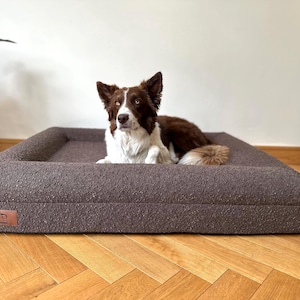 May include: A brown and white dog lies on a rectangular, brown dog bed. The bed has a textured fabric and a low, raised edge. The dog bed is on a wooden floor.