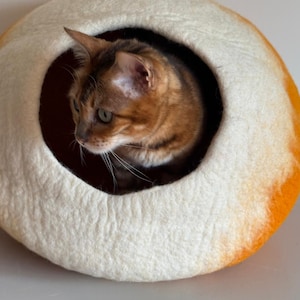 May include: A round, felted pet cave shaped like a loaf of bread with a cream-colored center and golden-brown crust edges. A brown and black tabby cat is positioned inside the circular opening of the soft, textured wool structure.