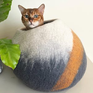 May include: A round, felted wool cat cave featuring a gradient color pattern that transitions from white at the top to dark gray at the base, accented by a diagonal stripe of warm orange wool. A brown tabby cat is resting inside the spherical structure.