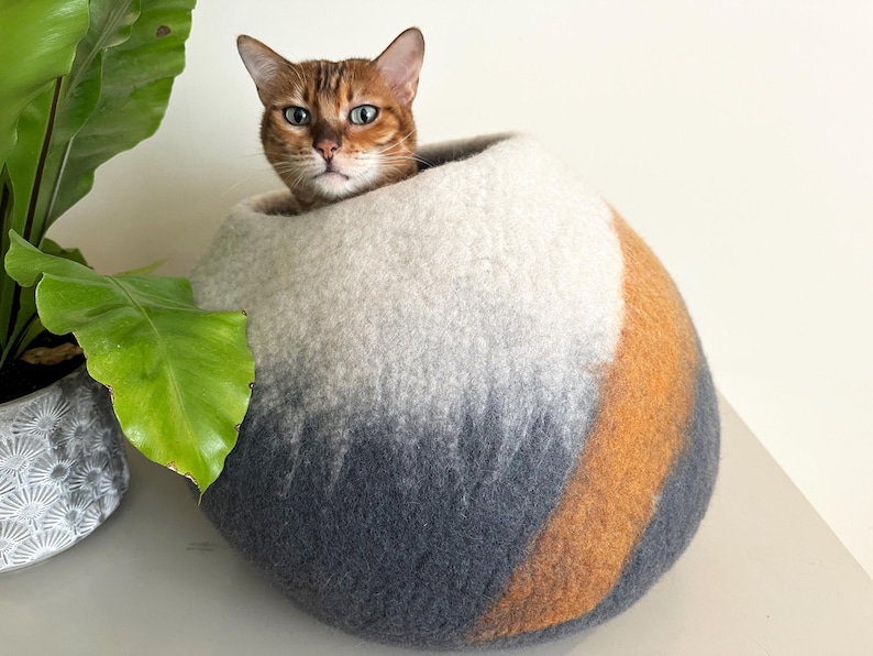 May include: A round, felted wool cat cave featuring a gradient color pattern that transitions from white at the top to dark gray at the base, accented by a diagonal stripe of warm orange wool. A brown tabby cat is resting inside the spherical structure.