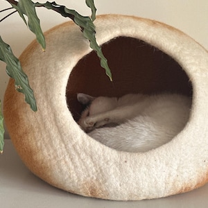 May include: A round, felted pet cave shaped like a loaf of bread with a tan crust and a cream-colored interior. A white cat is curled up inside the circular opening of the structure, which features a soft, textured surface throughout its exterior.